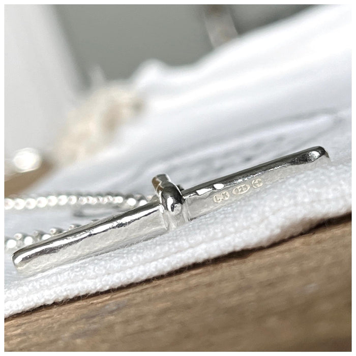 A silver T bar necklace with a hammered finish, laid on a belcher chain on a white surface.