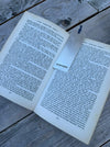 An open book spread on weathered grey wooden planks, with a white rectangular bookmark featuring the a large Hallmark.