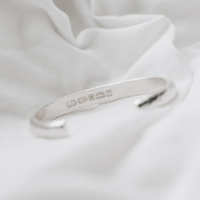 A silver chunky cuff bracelet with a hammered effect, displayed on a white fabric surface.