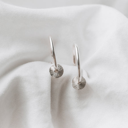 Sterling silver hoops with a textured disc on each, laying on a white linen background