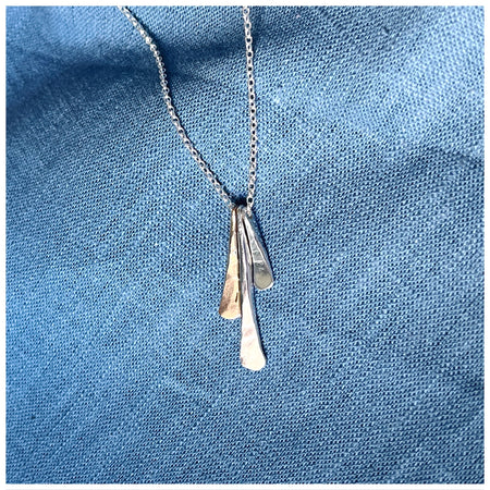 A silver pendant with three separate elements, in silver and gold hanging from a silver chain, displayed against a blue fabric background.