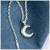 A silver crescent moon pendant on a chain, displayed against a blue textured background.