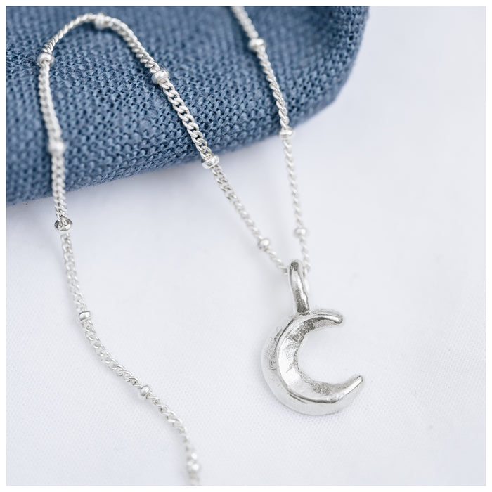 A silver crescent moon pendant attached to a small satellite chain, displayed against a blue textured background