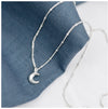 A silver crescent moon pendant attached to a small satellite chain, displayed against a blue textured background.