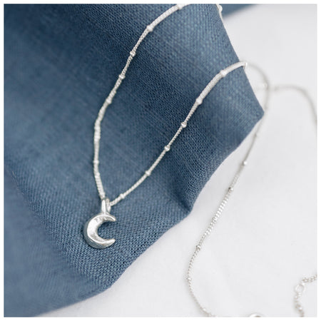 A silver crescent moon pendant attached to a small satellite chain, displayed against a blue textured background.