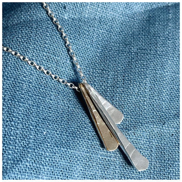 A silver pendant with three separate elements, in silver and gold hanging from a silver chain, displayed against a blue fabric background.