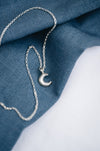 A silver crescent moon pendant on a chain, displayed against a blue textured background.