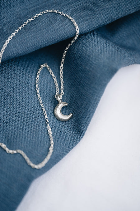 A silver crescent moon pendant on a chain, displayed against a blue textured background.
