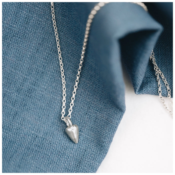 A silver heart-shaped pendant necklace with a delicate 1.7mm belcher chain, displayed on a blue fabric surface.