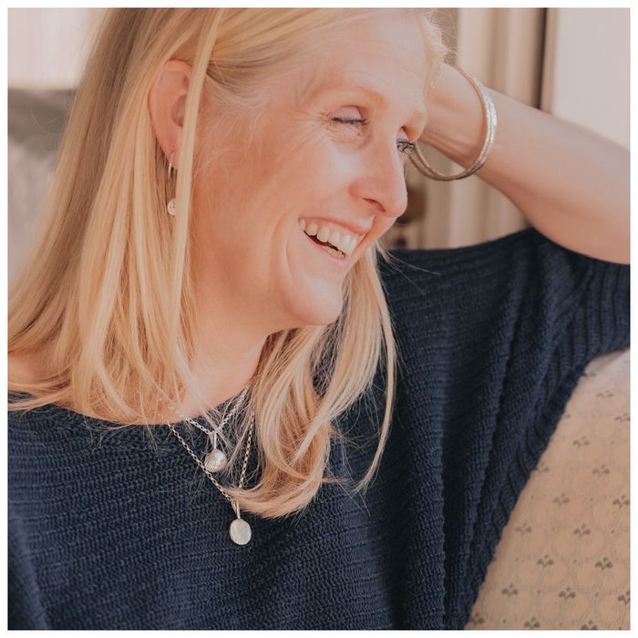 A 9ct gold full moon-shaped pendant necklace worn by a blonde lady in a navy jumper, stacked with a chunky shell necklace