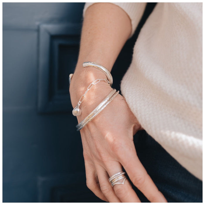 A silver chunky cuff bracelet with a hammered effect, stacked with a trace bracelet with a shell heart and a hammered bangle. 