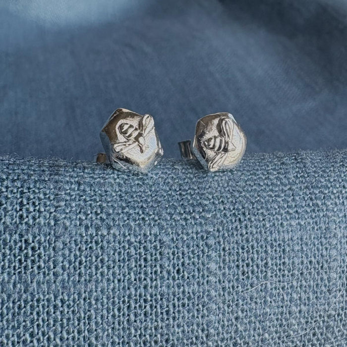 Sterling silver stud earrings with a bee on a piece of silver honeycomb sitting on a blue textured fabric background.