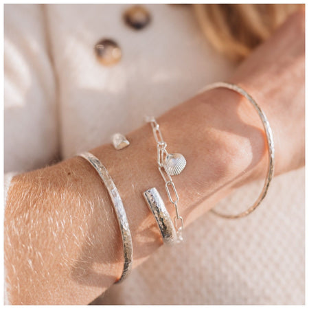 A silver chunky cuff bracelet with a hammered effect, stacked with a shell trace chain bracelet, a hammered bangle and a hammered bangle.